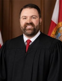 Portrait of Judge Taylor Bowman - The Judge is in a black robe with a white shirt and red tie and the State of Florida flag behind him